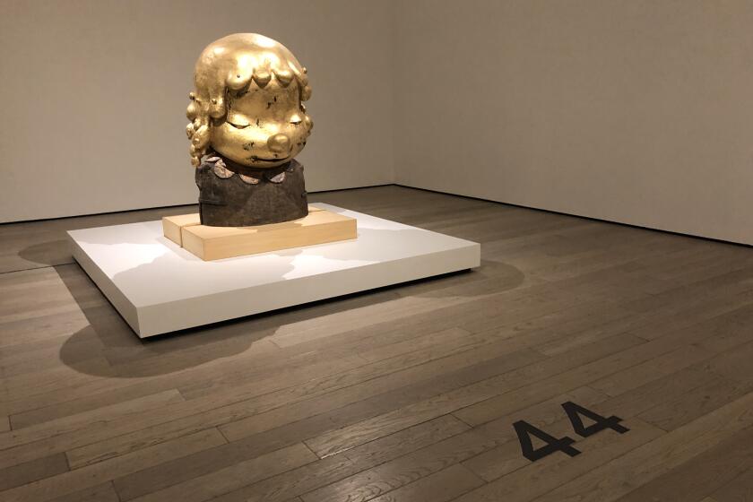 A golden sculpture of a feminine looking figure sits in a gallery. On the floor, a large number 44 marks where to stand
