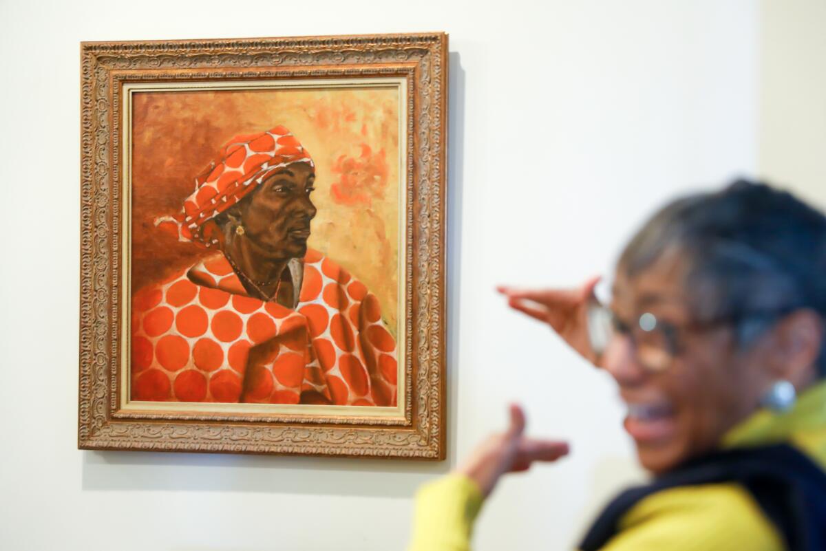 A woman gestures toward a hanging portrait.