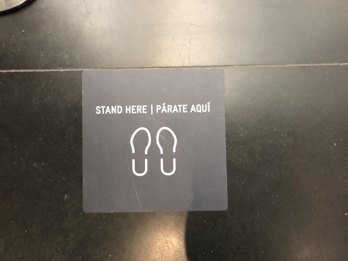 A floor marker shows the outline of two feet and the words "Stand Here / Párate Aquí"