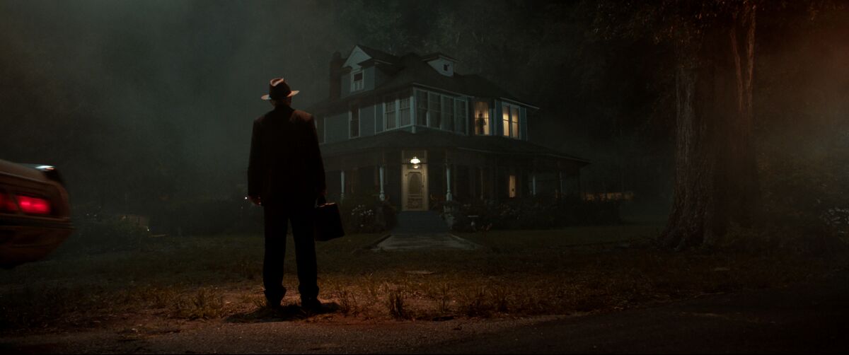 In the dark, a man in a fedora stands in front of a spooky-looking house.