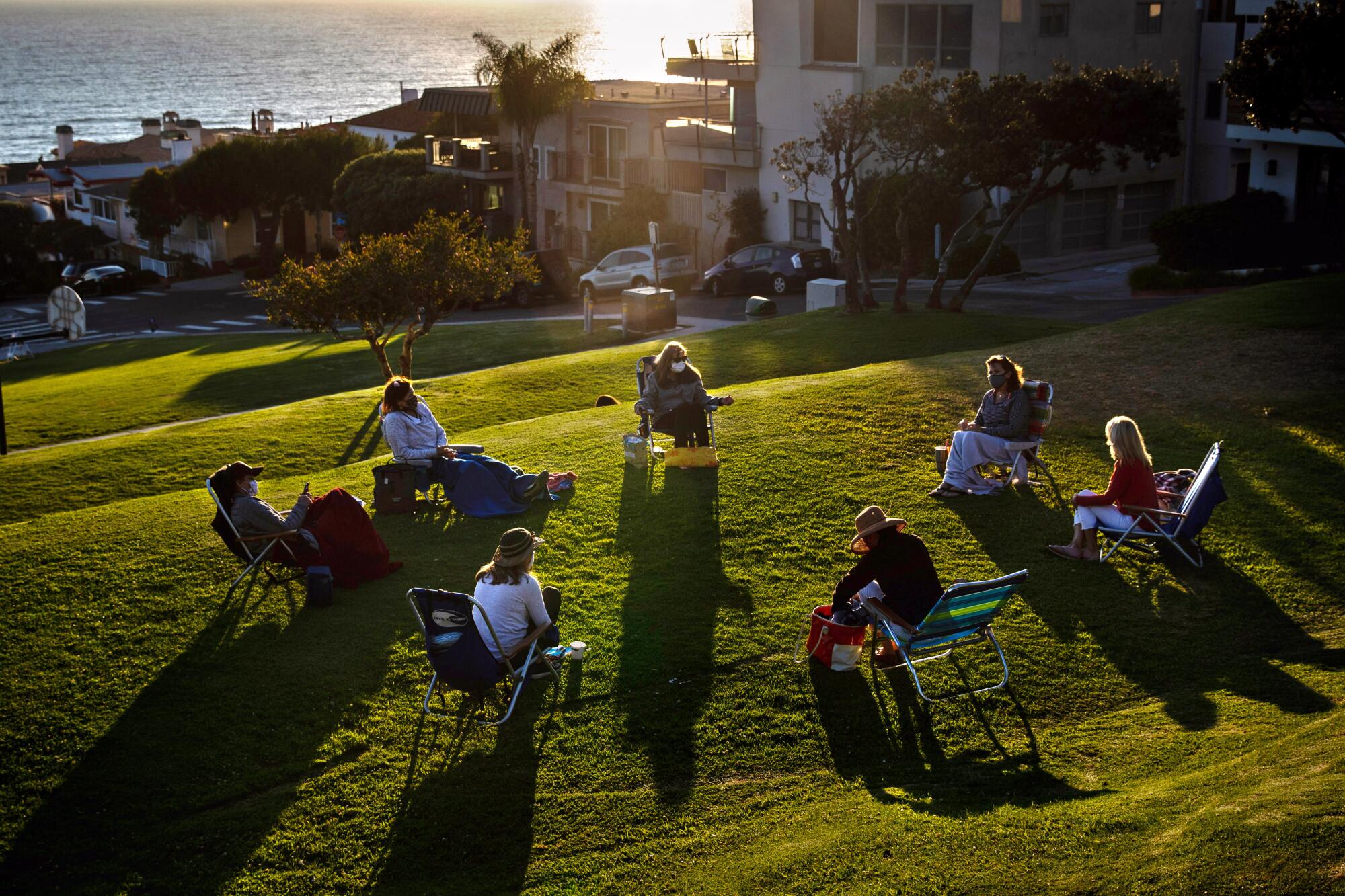 Seven people sit in lawn chairs arranged in a circle outdoors on a grassy sloped area, with the ocean in the background. 