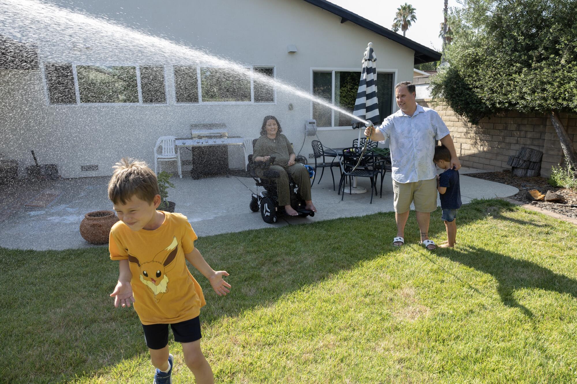 A father sprays a hose toward his son in a backyard of a suburban home. A child stands next to his dad; mother looks on. 