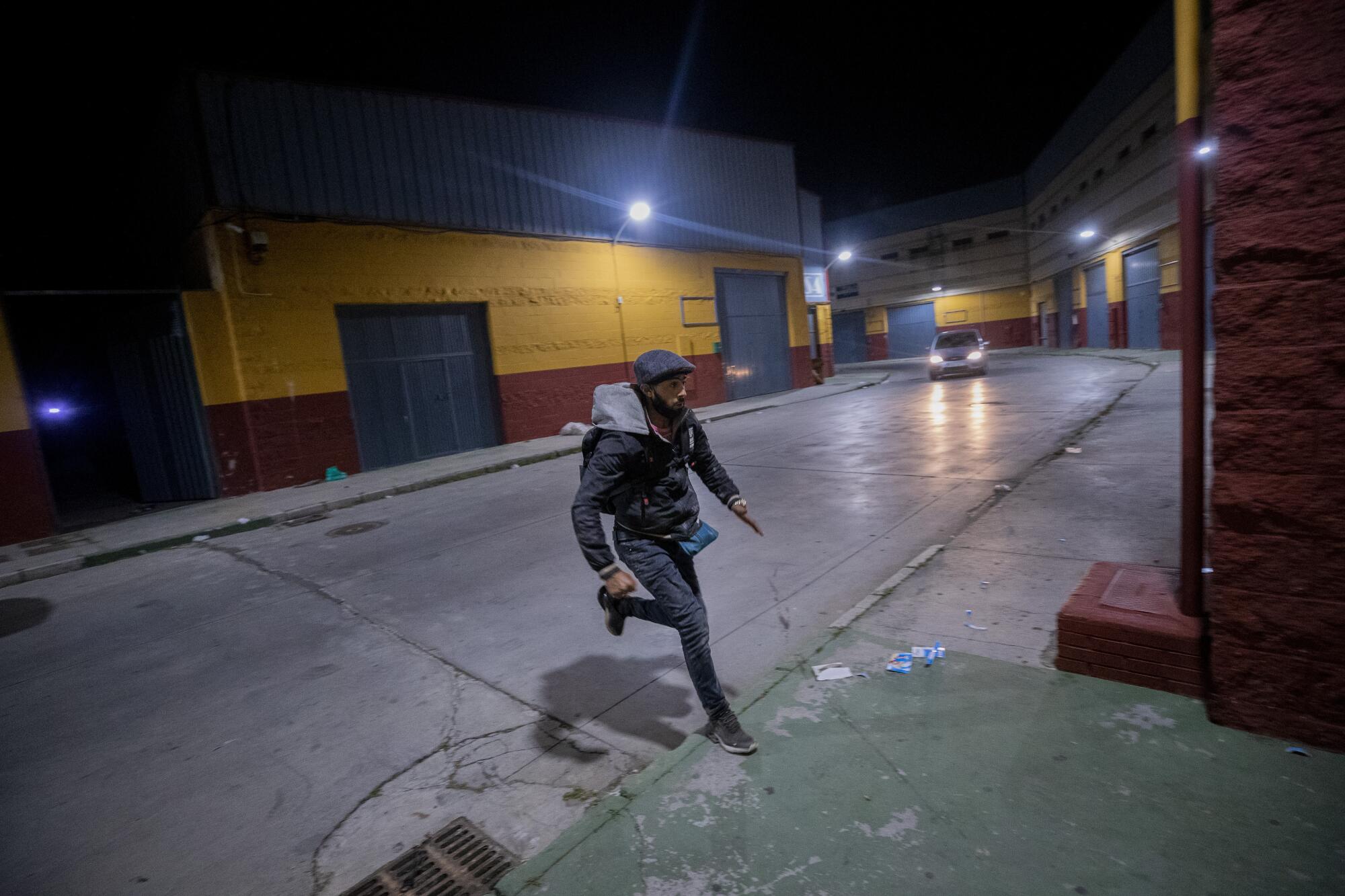 A man runs across a narrow street with a single car on it at night