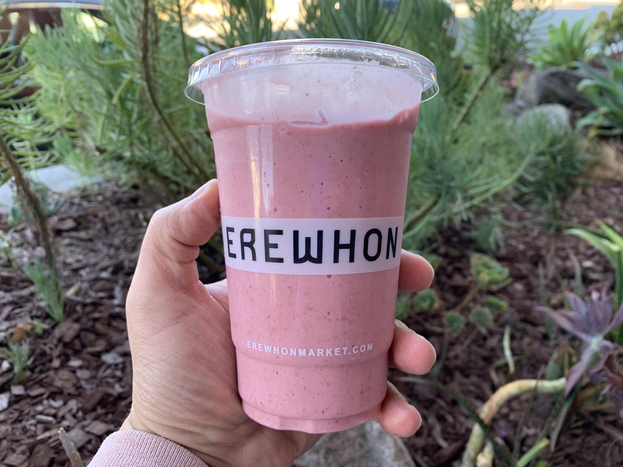 A hand holds a pink smoothie in a plastic cup that says Erewhon.
