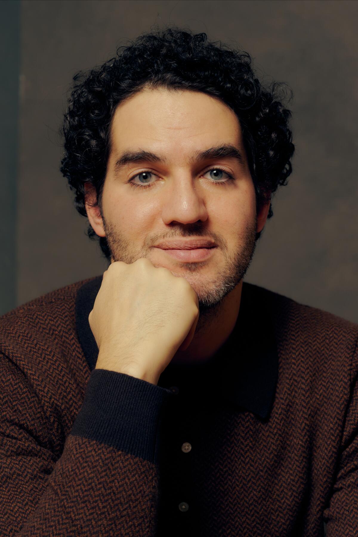 A man in a dark shirt, with dark curly hair, rests his chin on his hand and smiles slightly