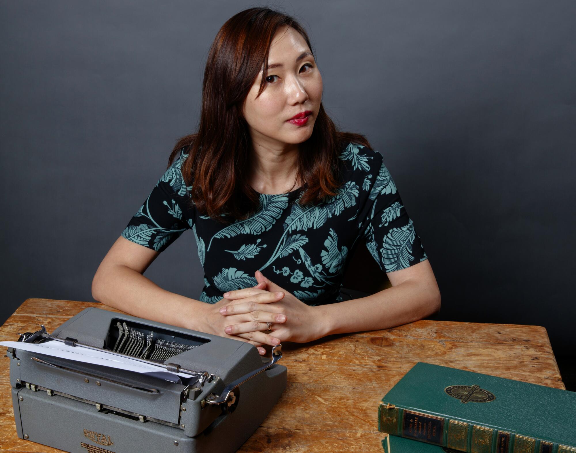 A woman with her hands clasped on a table with a typewriter and books.