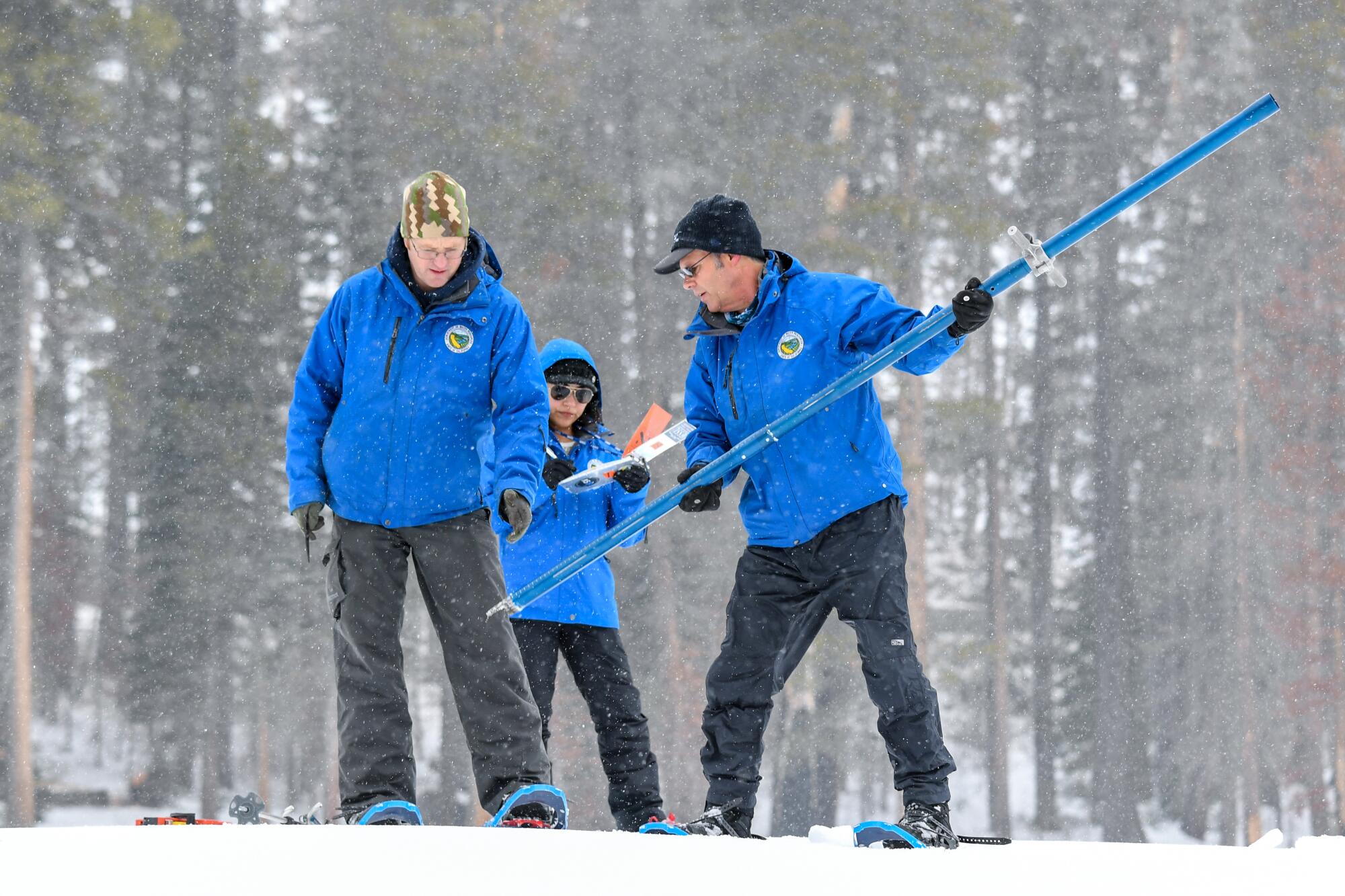A trio of workers wearing blue parkas and snow shoes prepare to measure snowpack with a long, tubular device. 