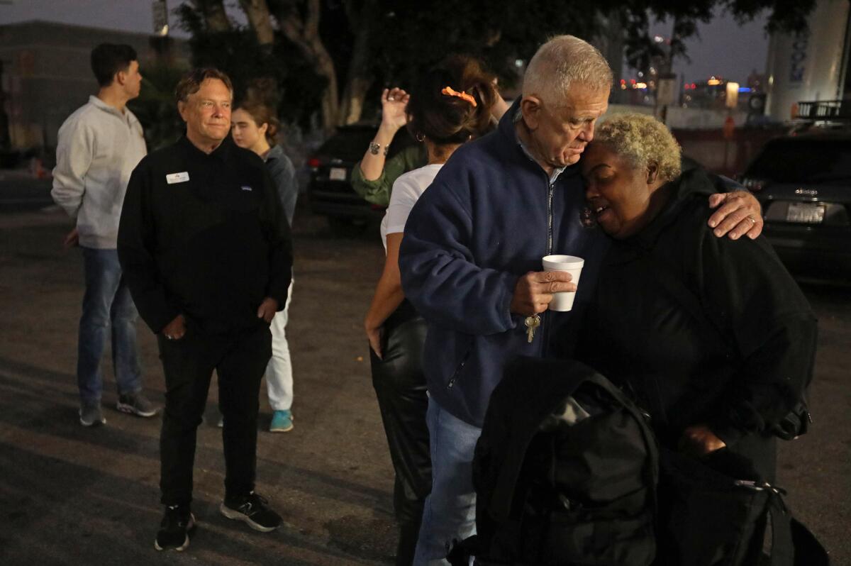 A man holding a cup gives a woman a hug while other people stand nearby. 