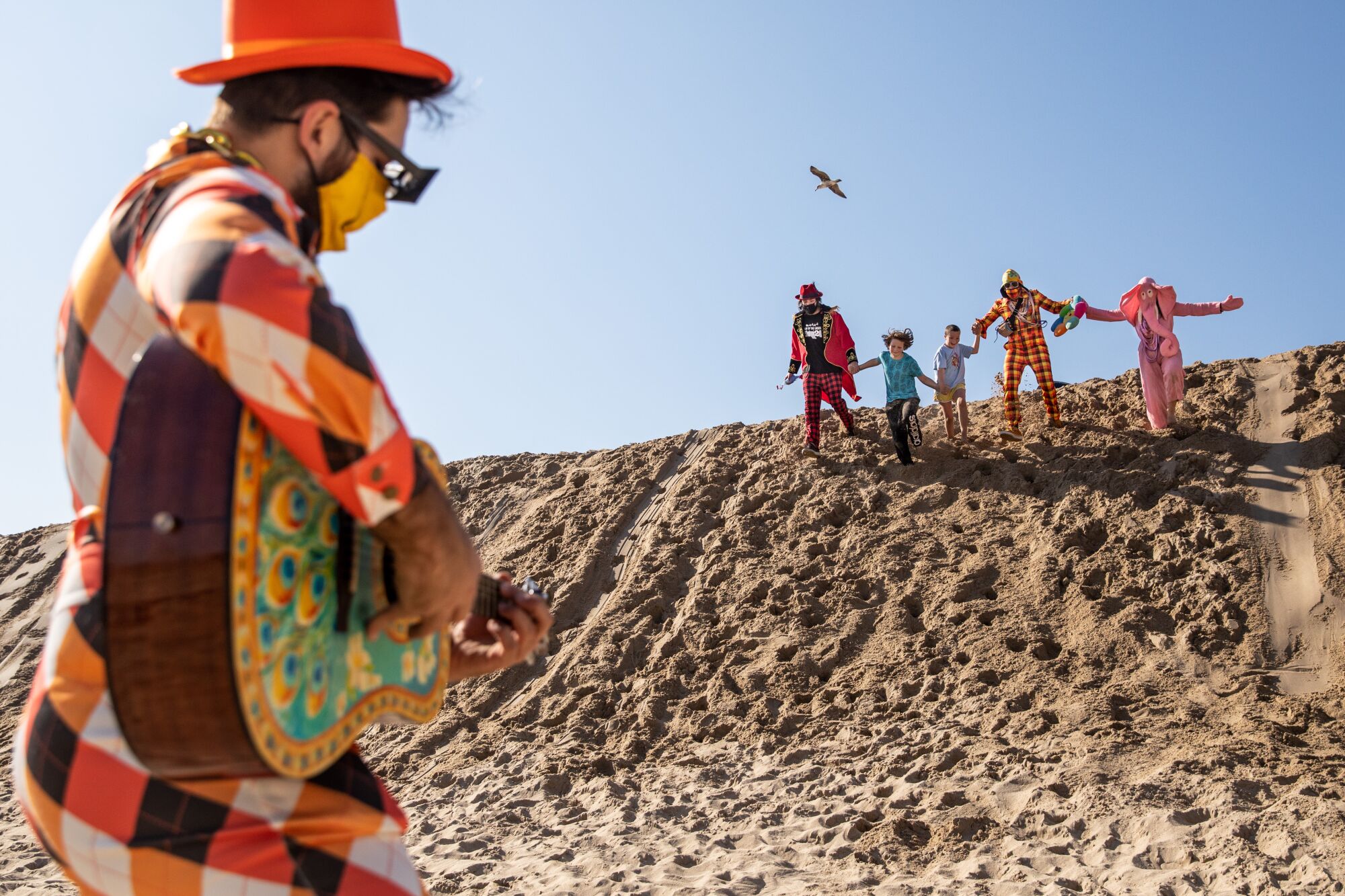 Daniel Drockton, in brightly colored outfit and hat, strums a guitar as people in costume descend a sand dune.