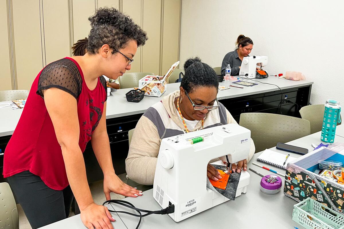 A person stands next to a person seated at a sewing machine. Another person is seated at a sewing machine behind them.