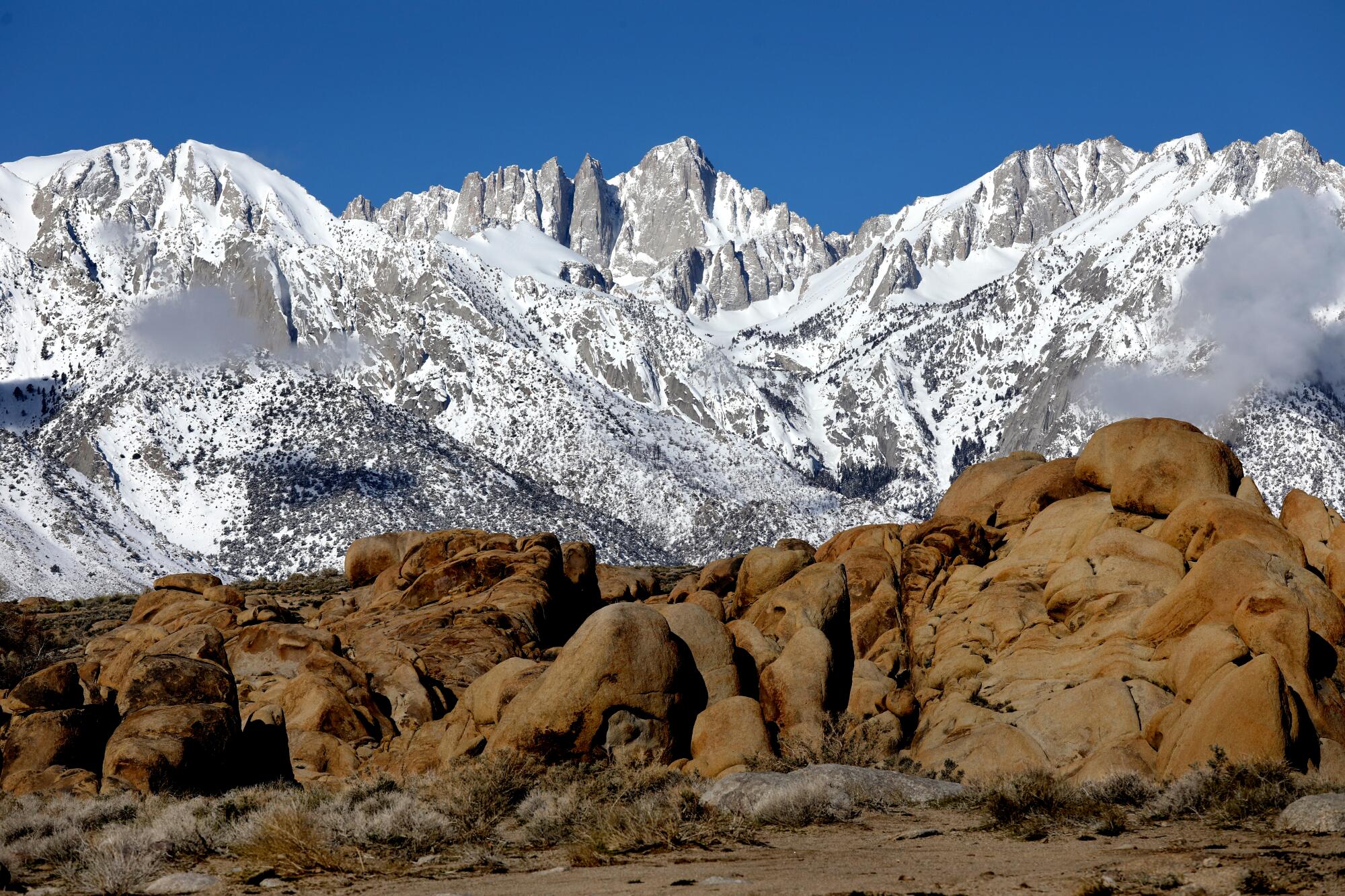 Snowy mountains on the horizon fill most of the frame. Dry, sand-stone rocks and land fills the foreground.