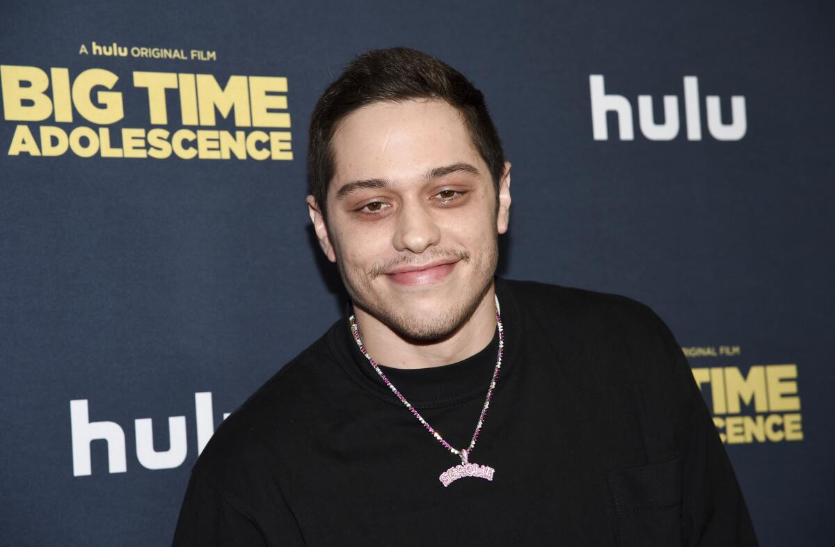Pete Davidson is smiling and posing while wearing a dark shirt and pink necklace