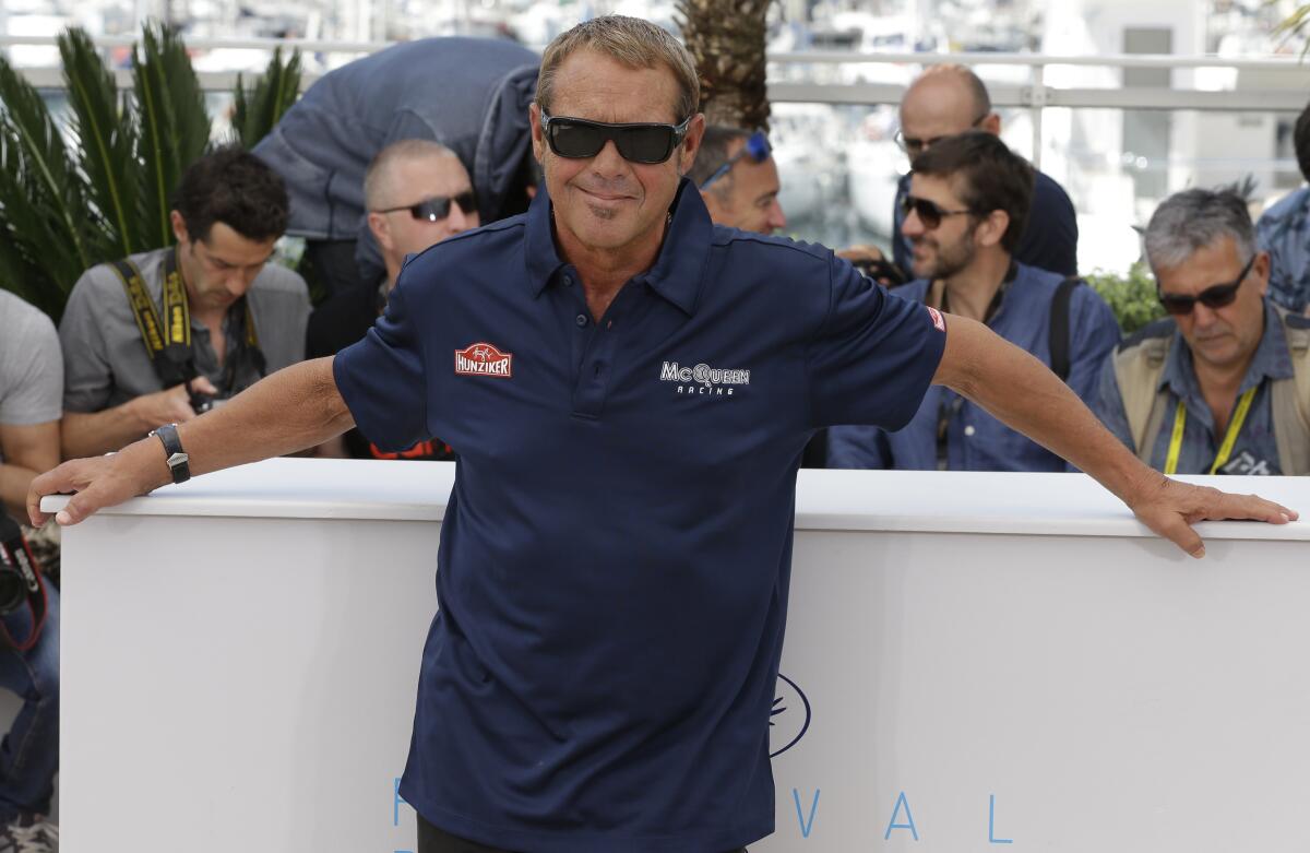 Chad McQueen in a blue shirt and shades posing in front of photographers with his arms spread wide on a tabletop behind him