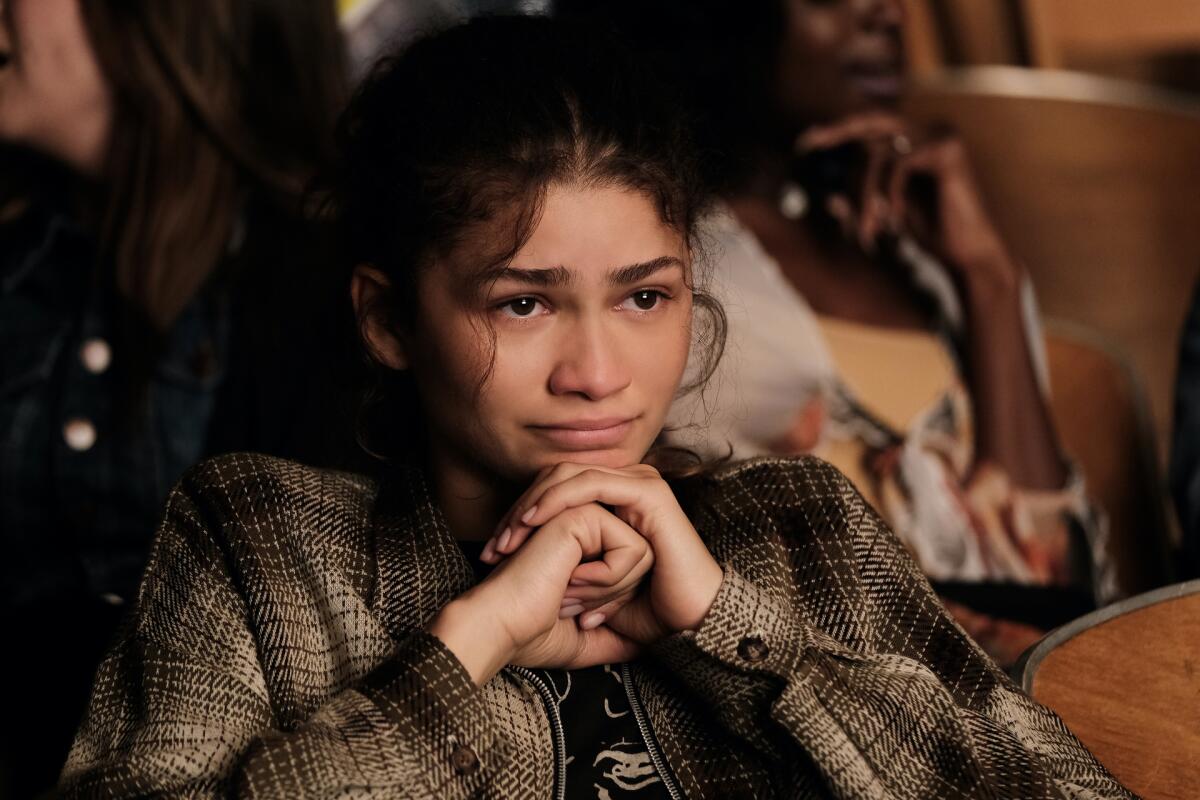 Zendaya clasps her hands under her chin and looks forward with sad eyes