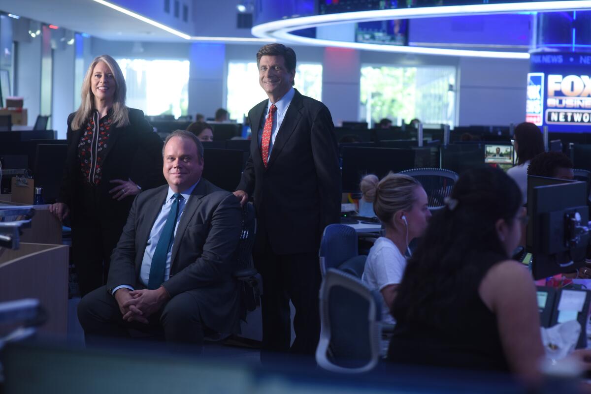 Three people pose for a photo amid others working in the Fox newsroom.