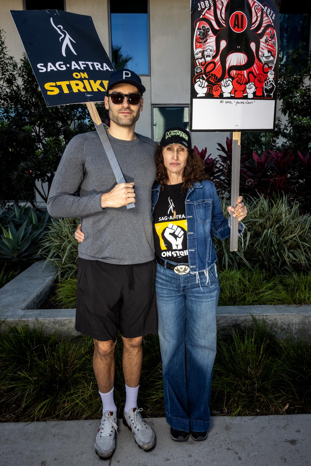 A man in a gray shirt and black shorts and a woman in a denim outfit pose together while holding picket signs.