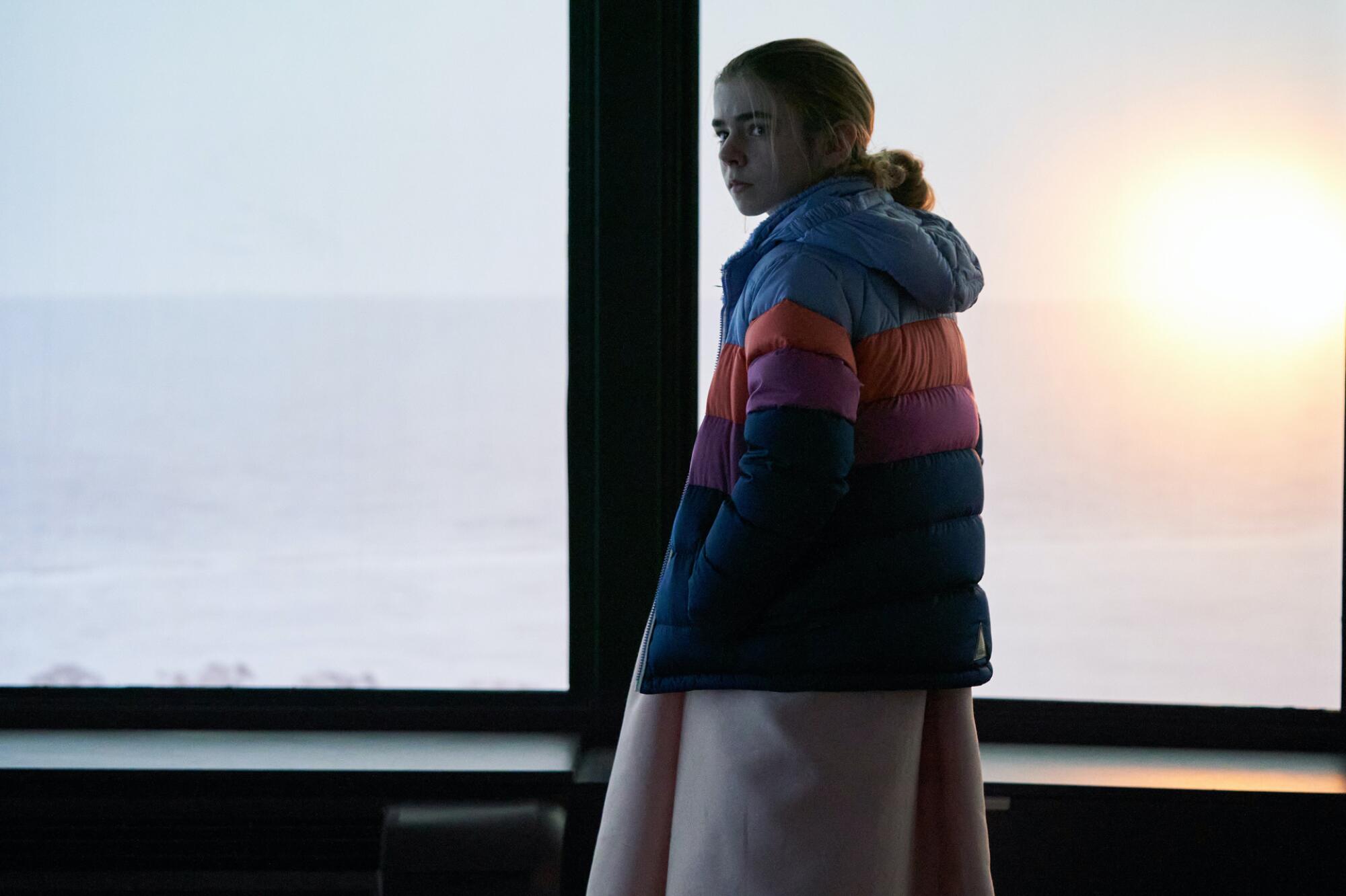 A pre-teen girl wears a coat indoors standing in front of a window in a scene from "Station Eleven."