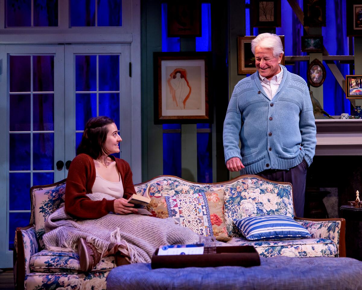 A man in a casual sweater stands next to a woman curled up on a couch on a stage set that looks like a middle class home