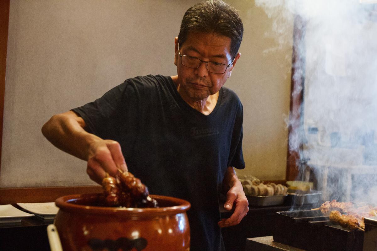Owner Shoji Ishikawa dunking yakitori and kushiyaki into a jar of tare next to a smoking grill