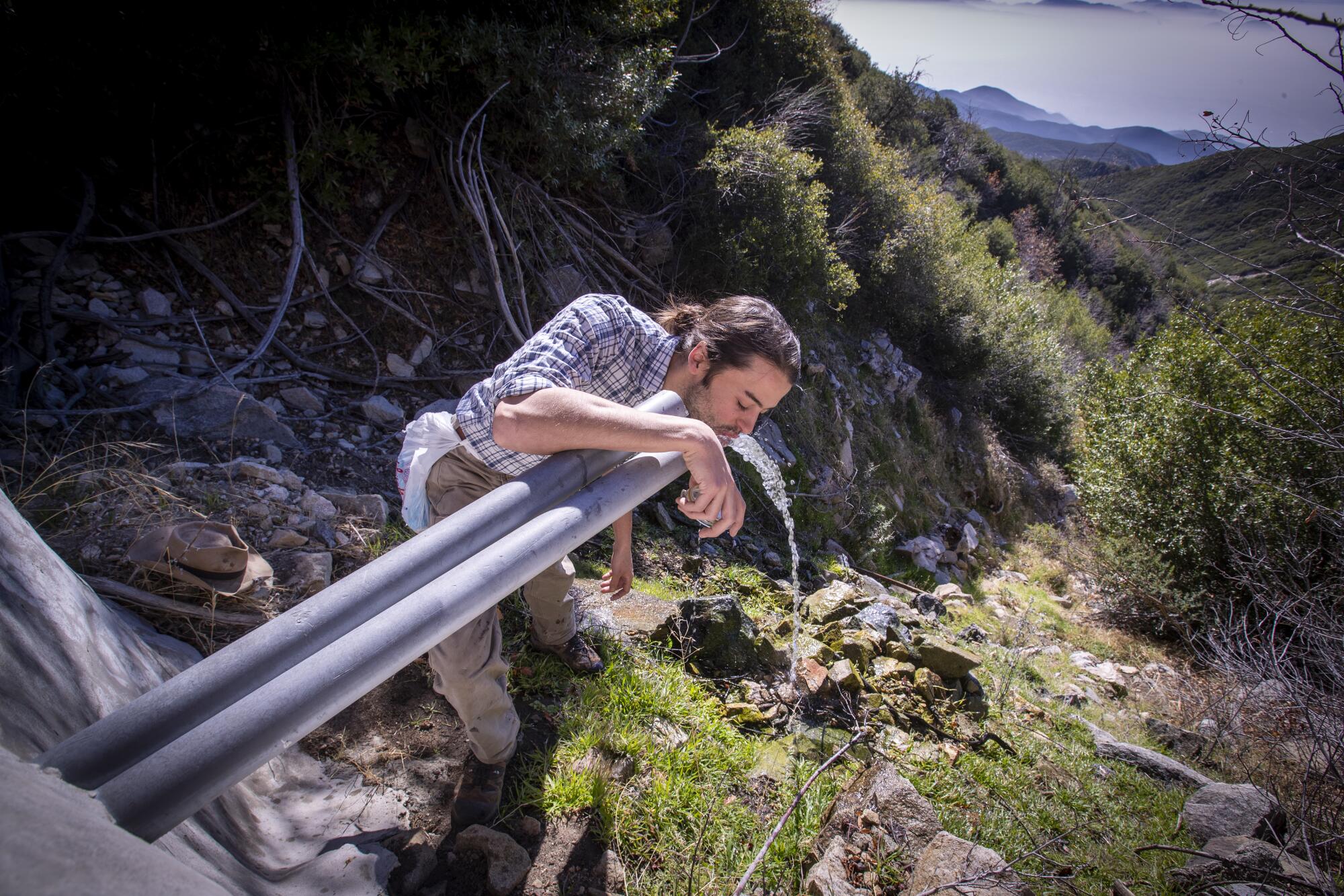 A man drinks water spilling from a pipe in a mountain forest.