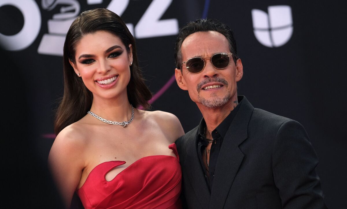 A woman with long brown hair in a red strapless dress smiles next to a man with black sunglasses and wearing a suit.