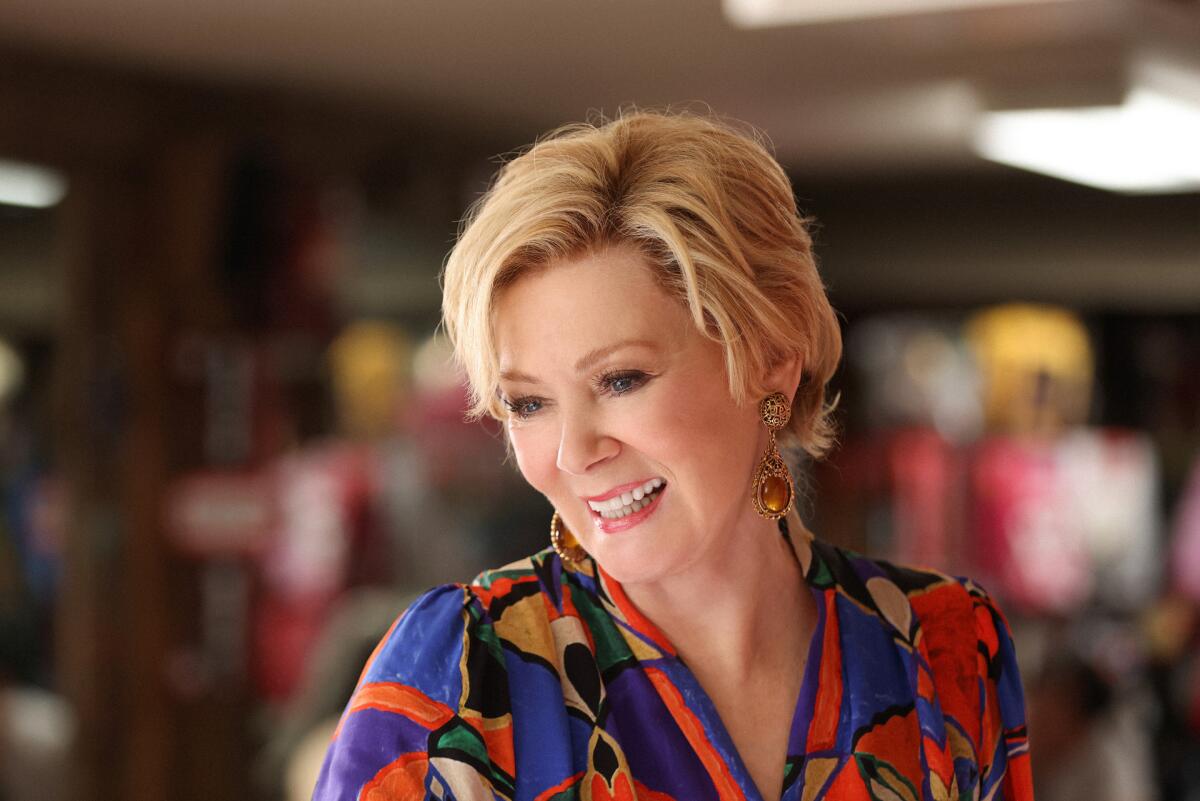 A woman with short blond hair in a multicolored blouse smiles as part of a TV show