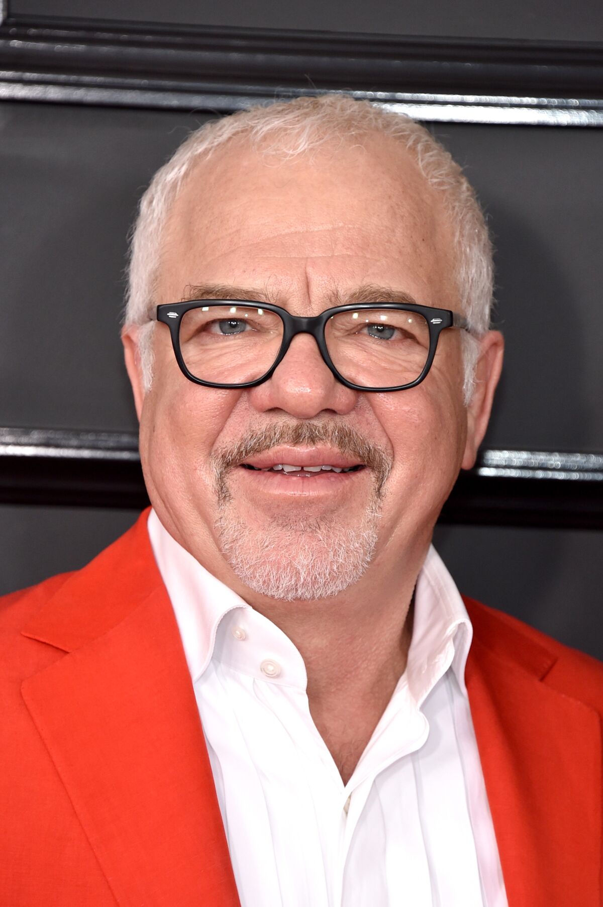 A man with a goatee in dark-rimmed glasses, an open white shirt and a red jacket.