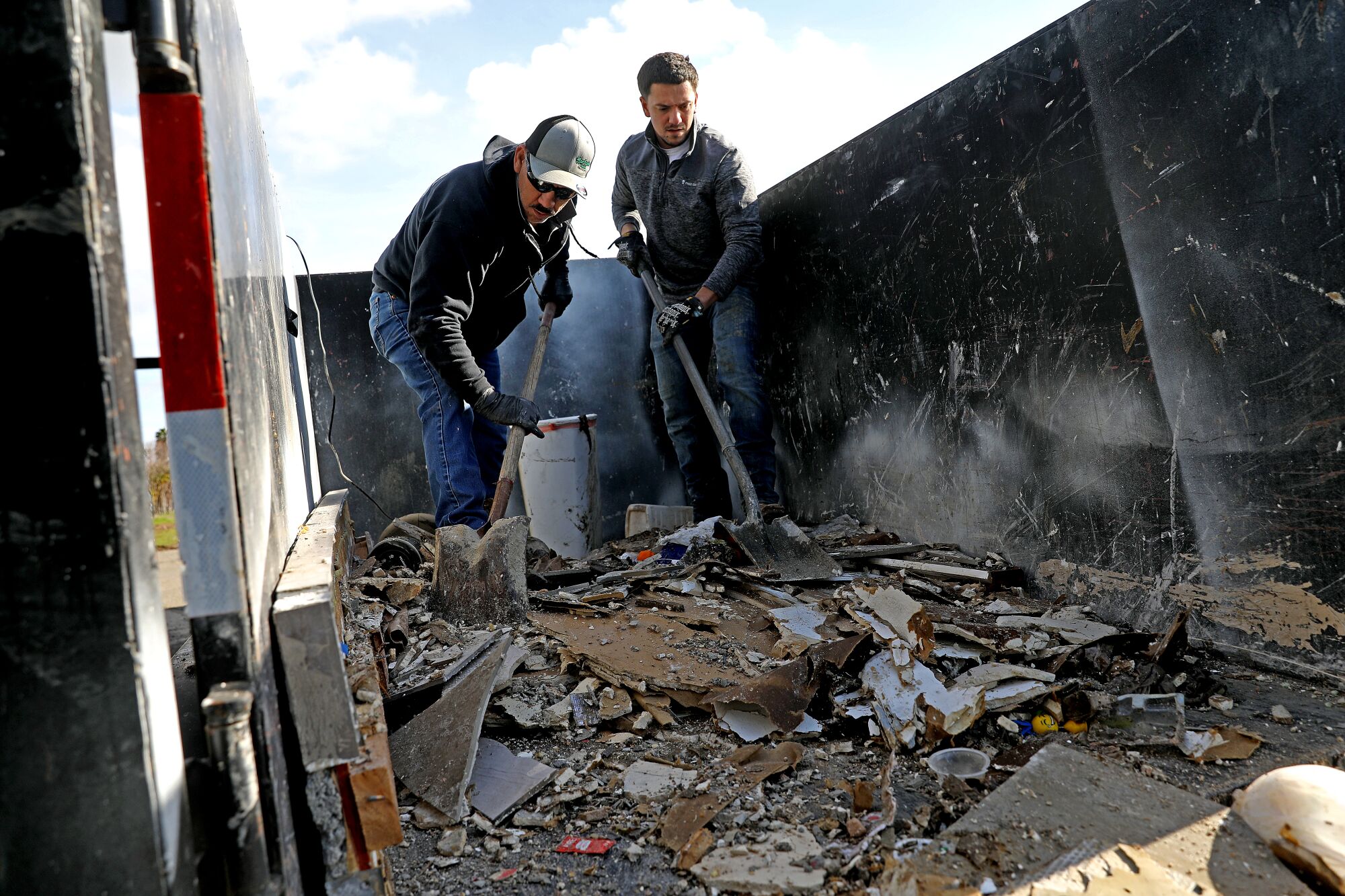 À l'aide de pelles, deux hommes déplacent des morceaux de débris d'inondation d'une remorque dans une poubelle.