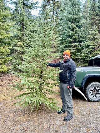 A man holds an evergreen tree in one hand and a saw in the other.