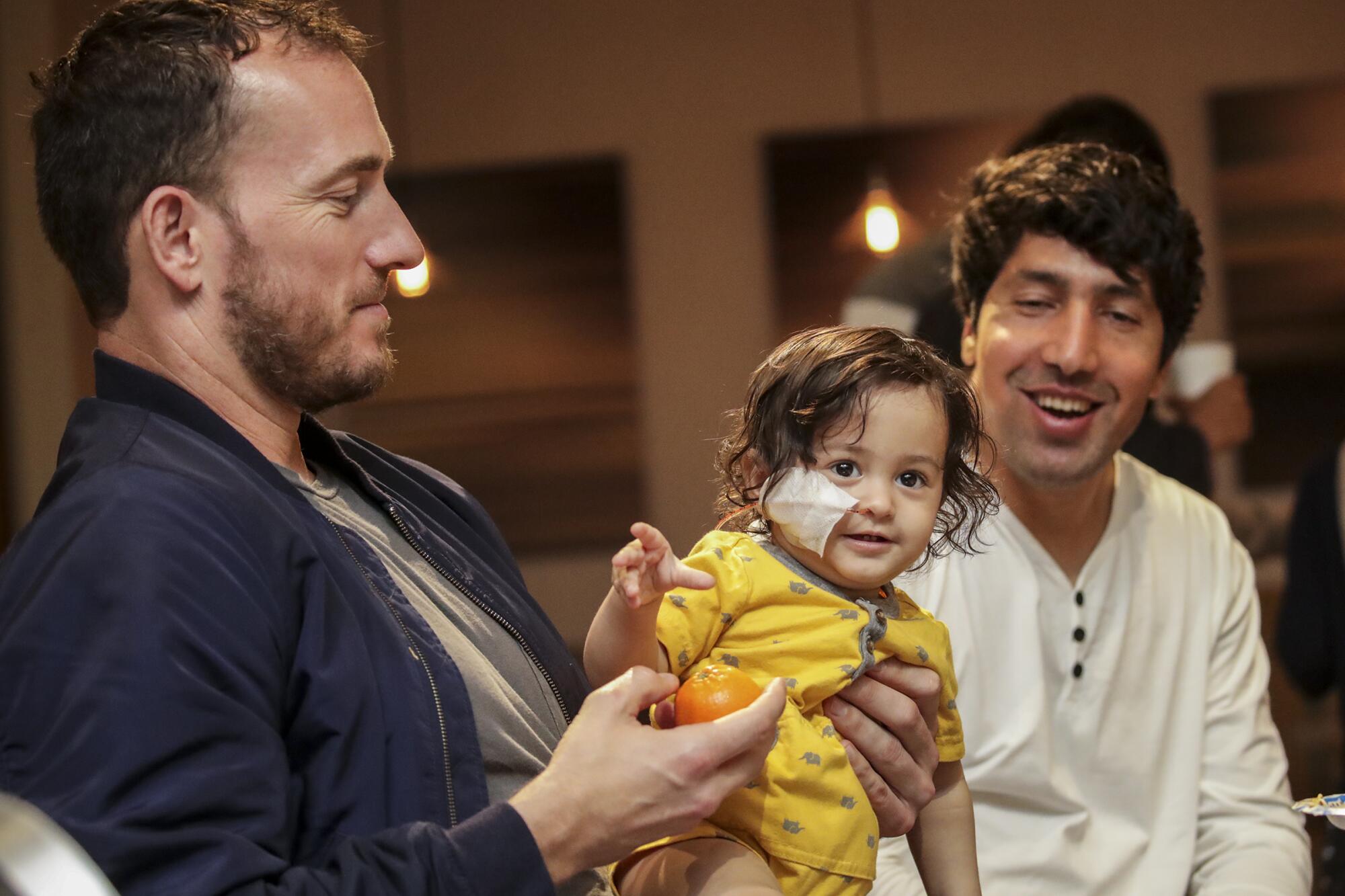 A man holds a child as another man watch.