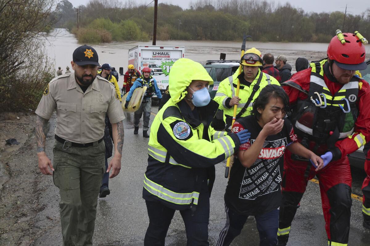 People in reflective gear and helmets walk with a weeping woman. In the background is a flooded roadway.