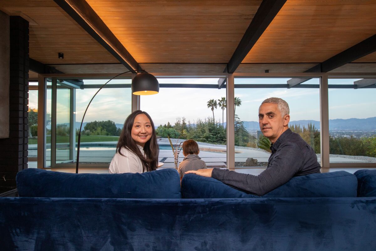 A couple seated on a sofa look over their shoulders at the camera, a boy and glass windows with a great view before them.