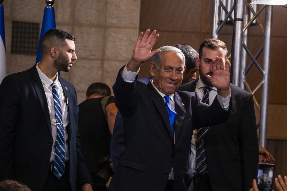 A man in a dark suit and blue tie has his hands up in acknowledgment, flanked by men in dark suits and striped ties
