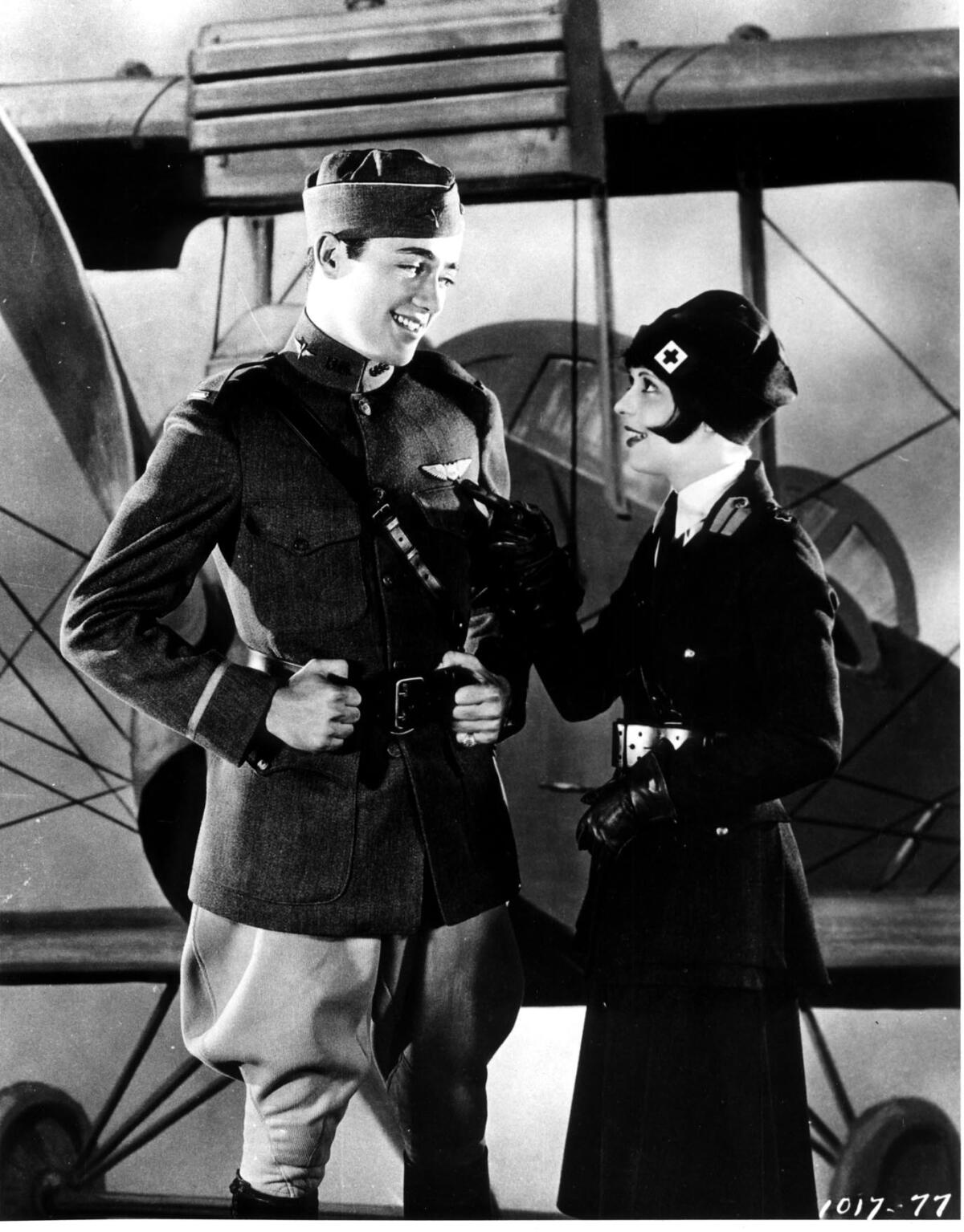 A man in uniform talks to a woman standing next to a 1920s airplane.