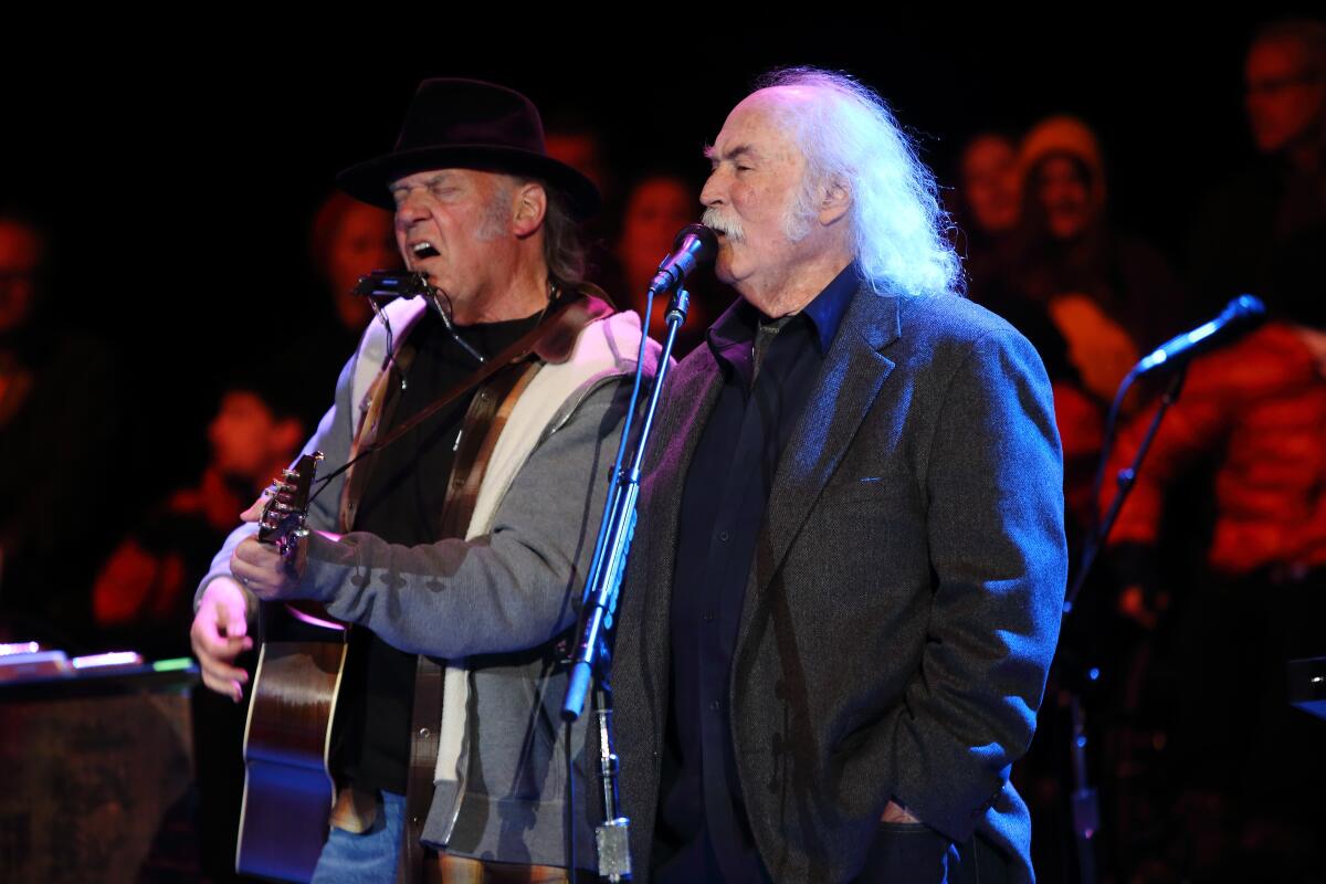 Two men, one in a jacket holding a guitar and another in a suit, singing at mics on a stage.