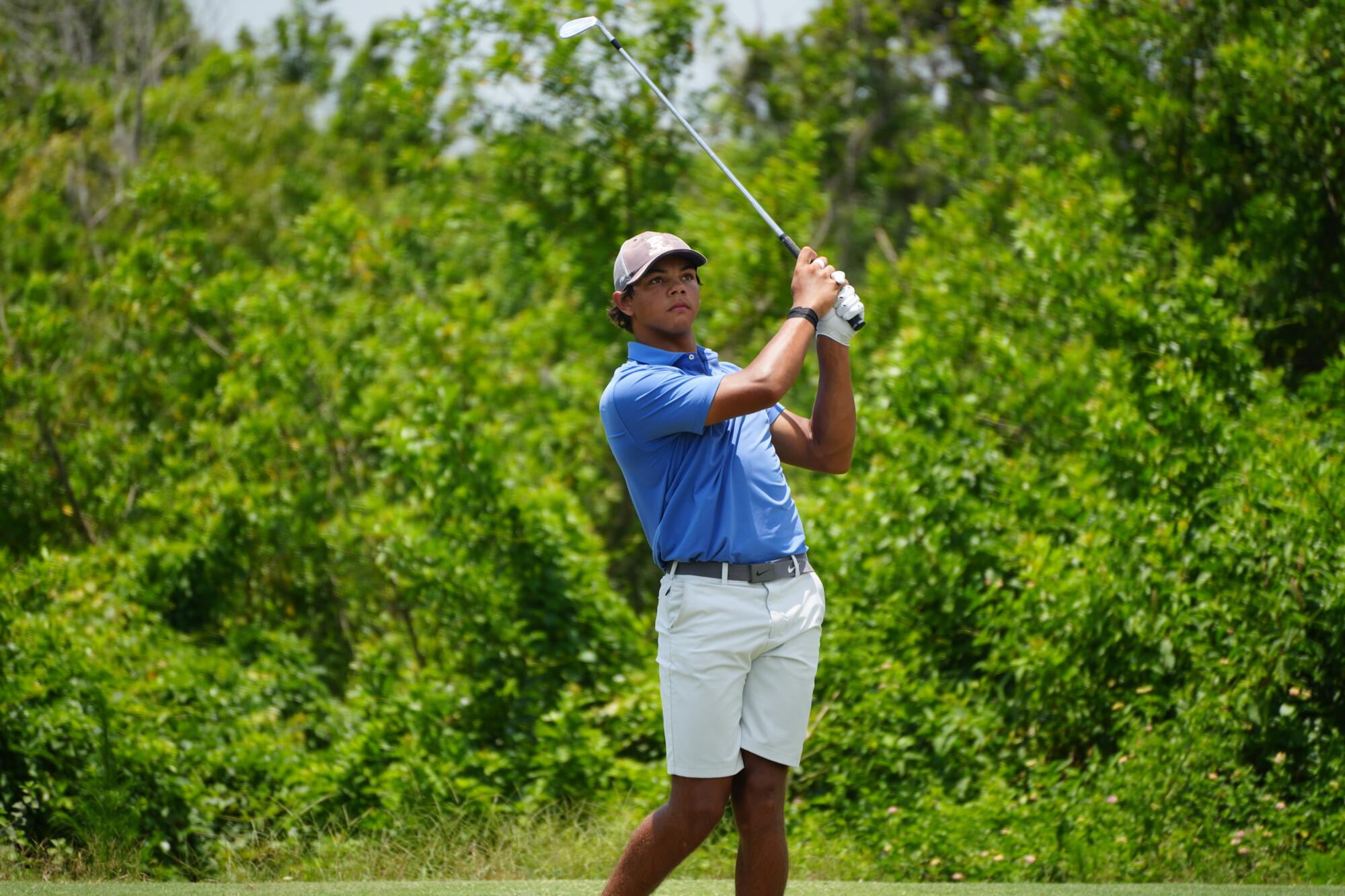 Charlie Woods wears a white hat, light blue shirt and white shorts and watches his shot, holding his club above his shoulder