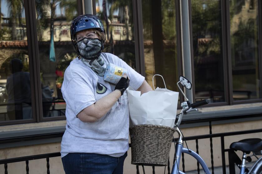 RIVERSIDE, CALIFORNIA - APRIL 3, 2020: Wearing a cat mask, Chani Beeman of Riverside is pleasantly surprised when she finds a roll of toilet paper inside her takeout food bag which she just picked up at Mario's Place during the coronavirus pandemic on April 3, 2020 in downtown Riverside, California. She rode her bike to the restaurant from her home about 1 mile away. Mario's is open for takeout only. Locals are ordering online and picking up at the front door Monday-Saturday 5pm - 8pm. The restaurant is located across the street from the historic Mission Inn, but the inn is now closed because of the pandemic.(Gina Ferazzi/Los Angeles Times)