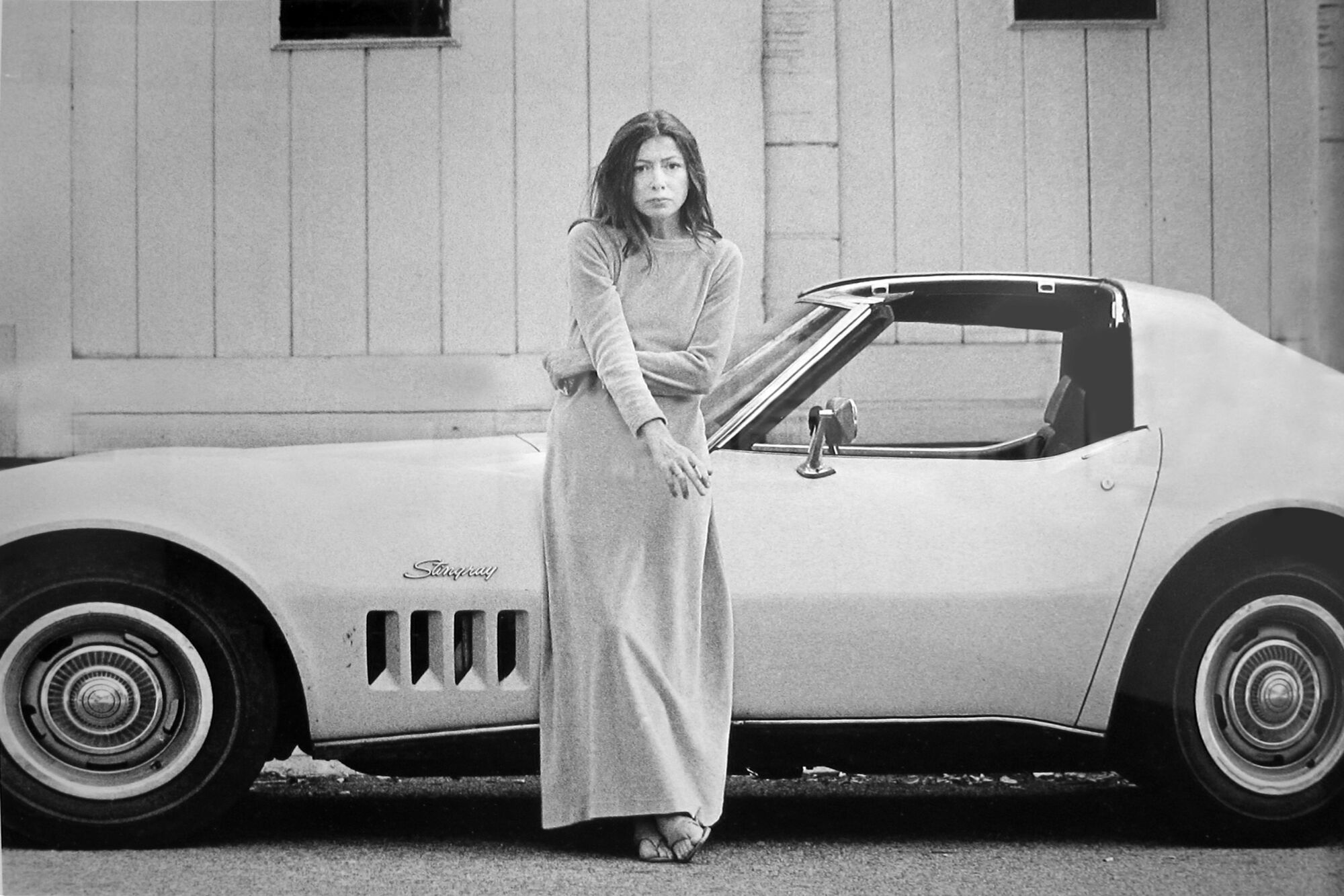A portrait of Joan Didion standing with a cigarette in front of her Corvette Stingray in 1968.