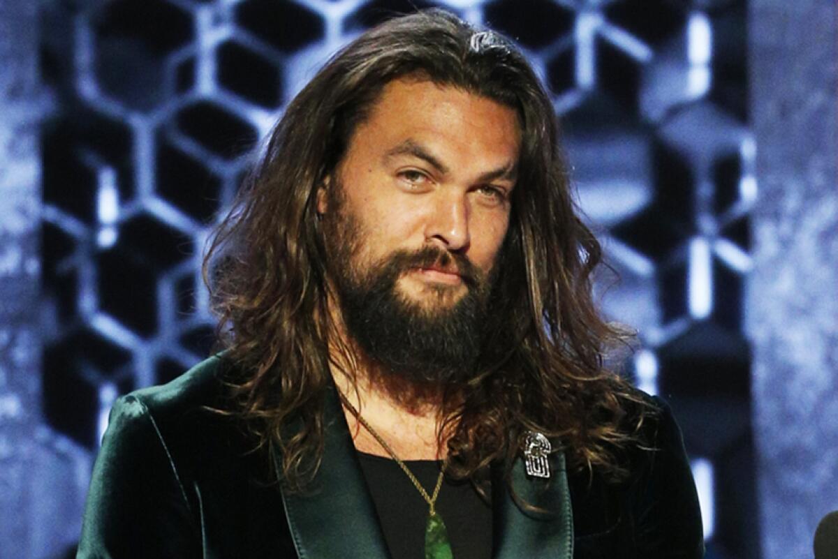 Jason Momoa poses with disheveled hair in a green velvet blazer and a green pendant necklace. 