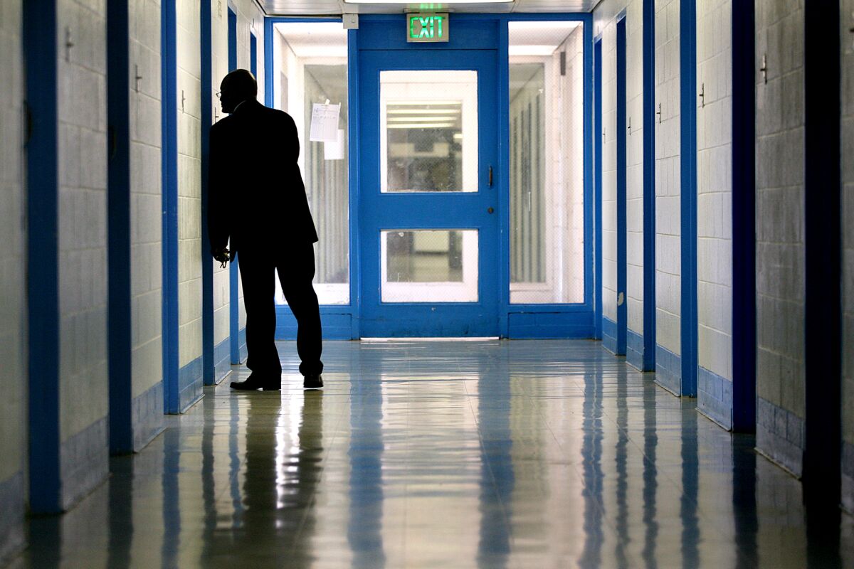 A frame of a darkened hallway with a row of doors. A silhouette of a man in a suit peers into a doorframe at right.