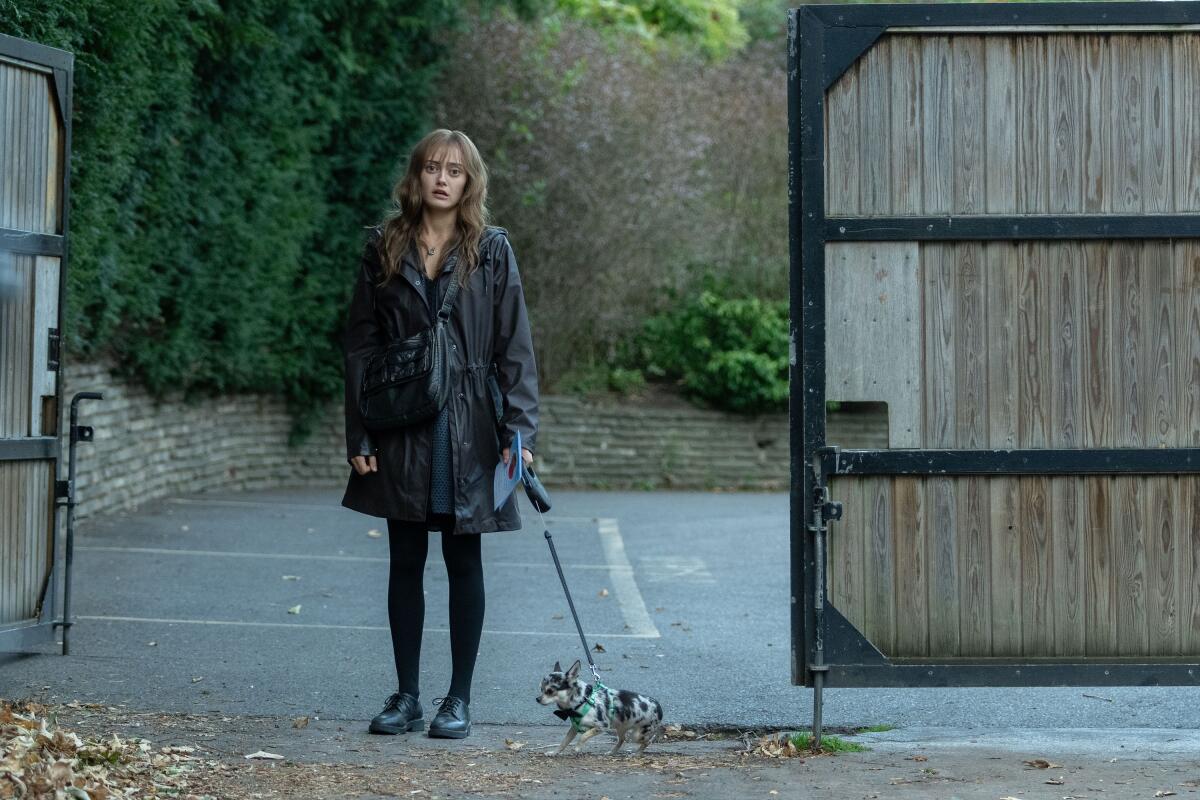 A woman in a dark coat, leggings and shoes stands at an open gate with a small dog on a leash.