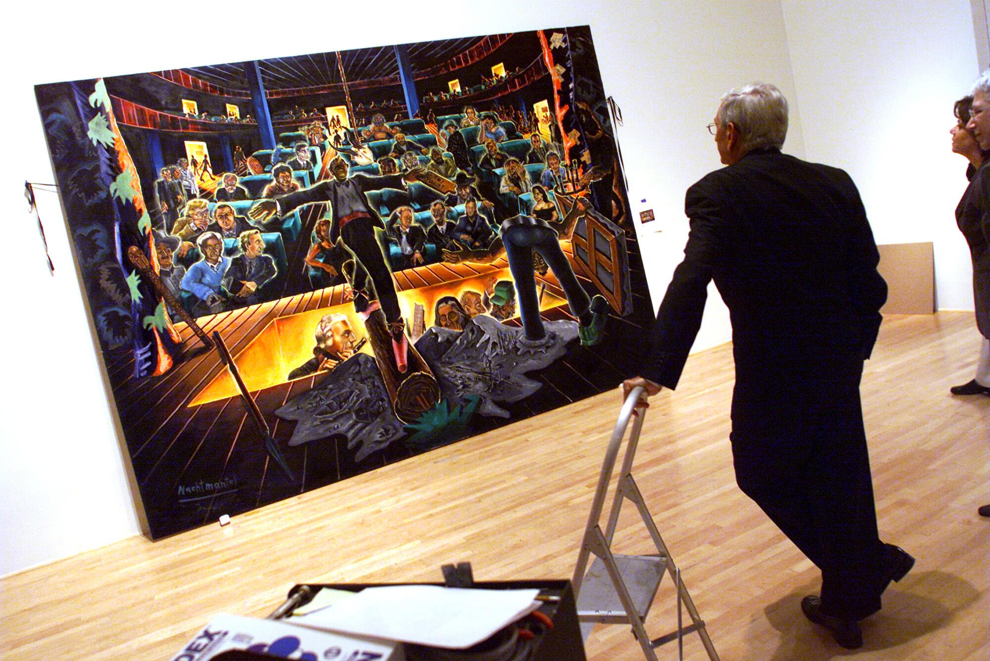 Eli Broad leans with one hand on a stepladder as he looks at a large painting.