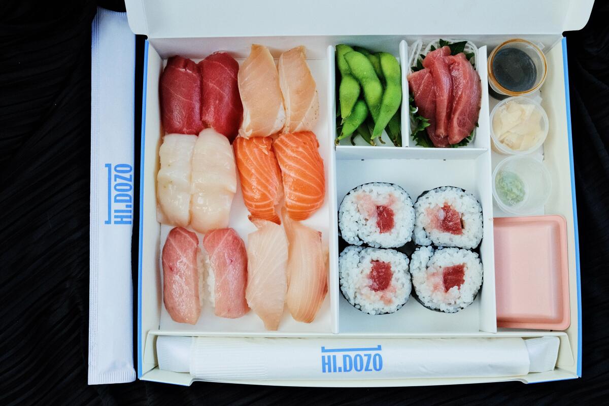 A white paper box of sushi featuring 12 pieces of nigiri, four of maki and sashimi with edamame.