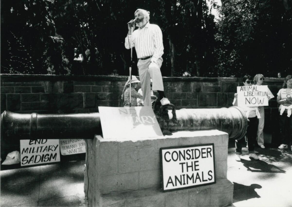 Elliot Katz stands on a vintage cannon surrounded by signs reading "Consider the animals" and "End military sadism."