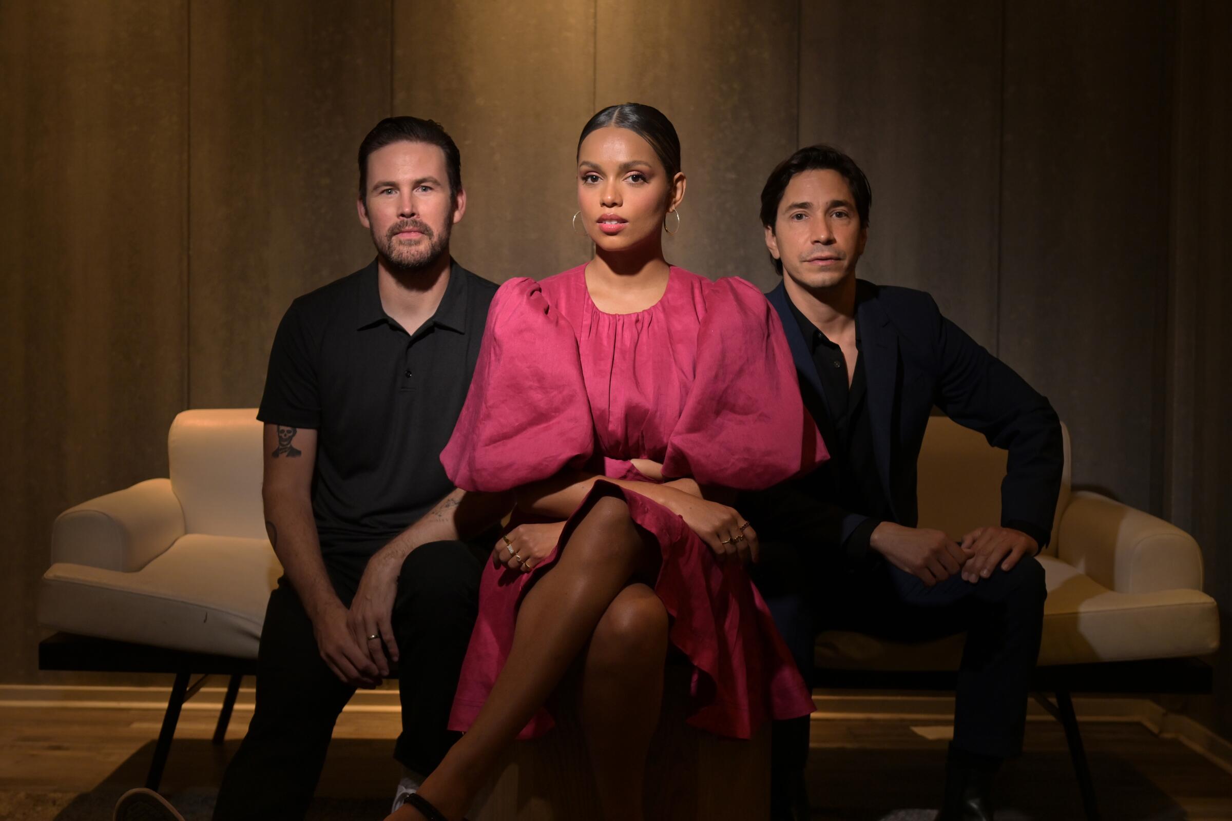 In a portrait in a subtly lit room, a woman in a red dress is flanked by two men in black.