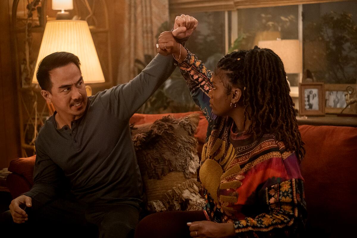 A man and a woman seated on a couch raise their arms to each other, their wrists touching.
