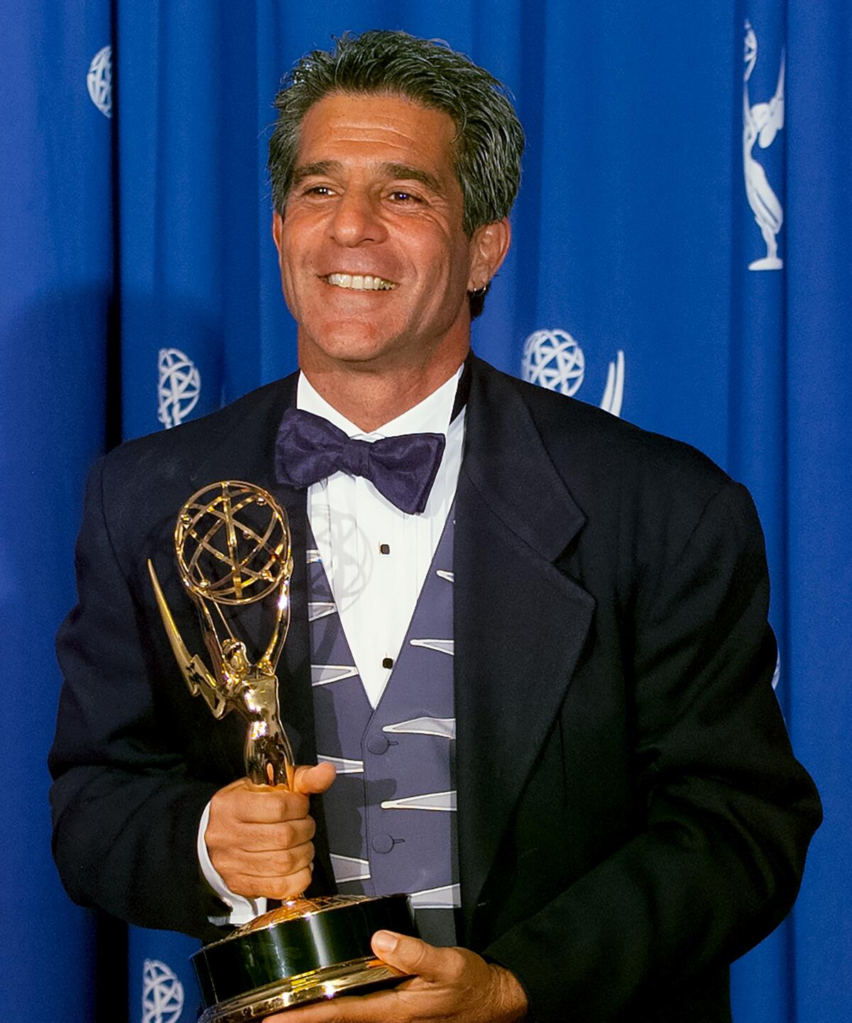 Michael Lembeck, in a tux with a purple vest and bow tie, holds an Emmy. 