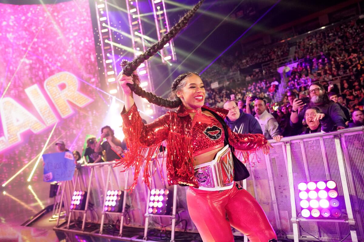 Bianca Belair walks in front of a crowd, smiling and twirling her hair.
