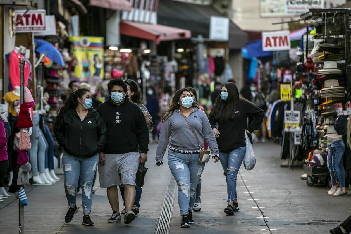 People in masks walk past clothing and other wares displayed in front of stores.