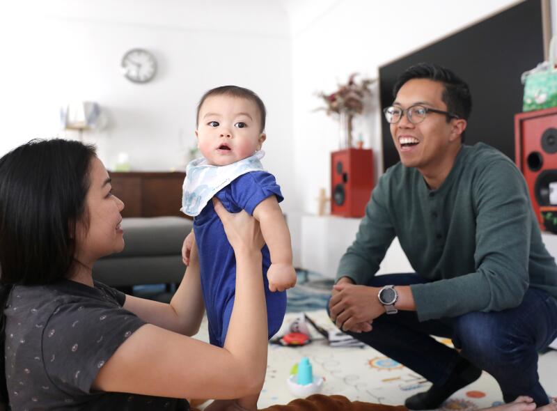 A couple playing with a baby inside a living room. One parent is lifting the infant up as the child looks towards camera